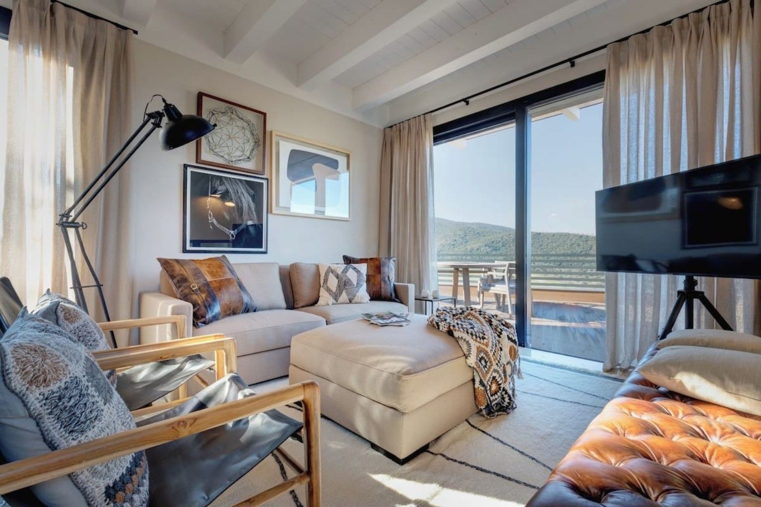 Ein modernes Wohnzimmer mit einer Couch, Hocker und einem Fernseher. Ein großes Fenster bietet Blick ins Freie.