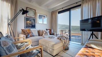 Ein modernes Wohnzimmer mit einer Couch, Hocker und einem Fernseher. Ein großes Fenster bietet Blick ins Freie.