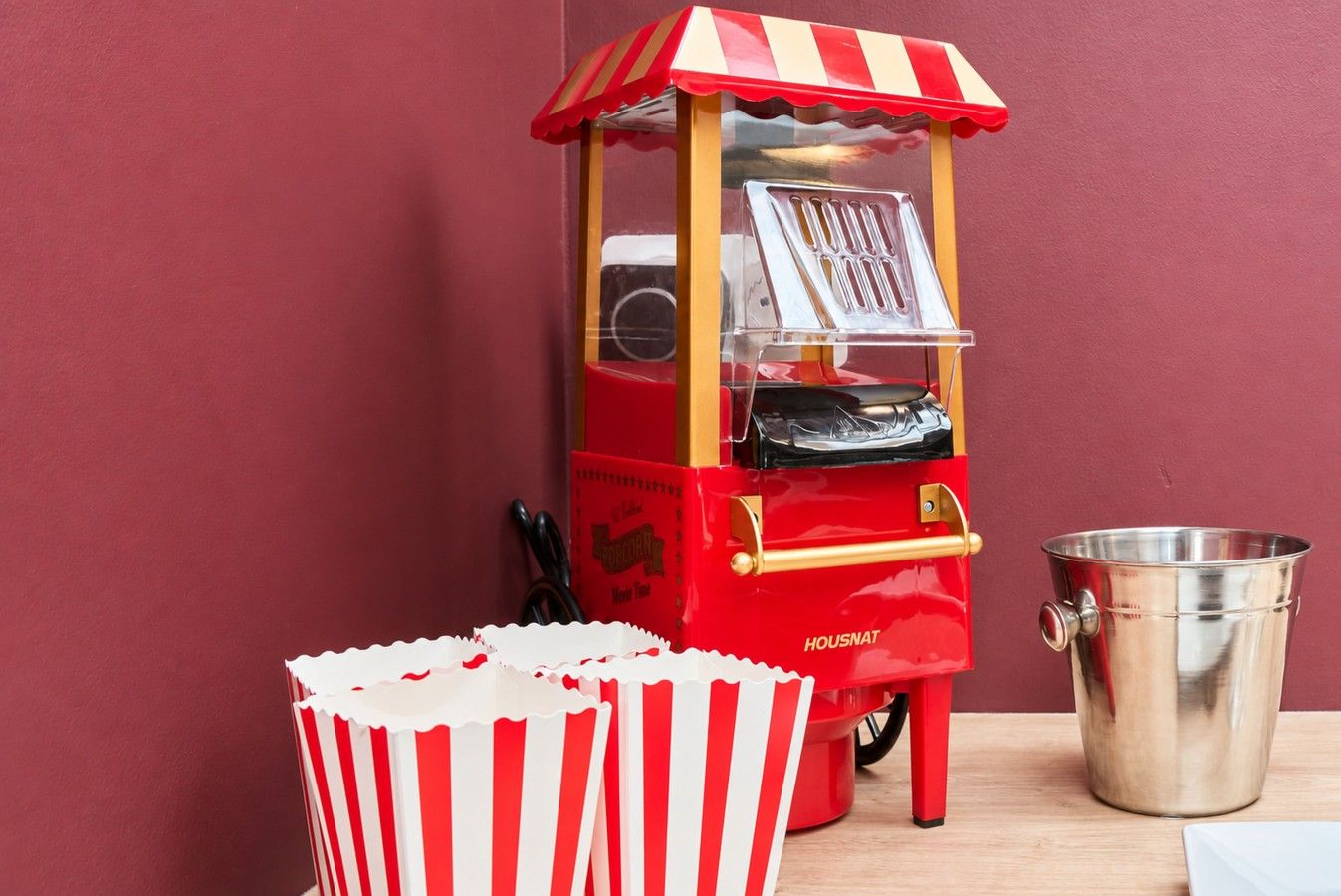 Máquina de palomitas roja y blanca con una ventana transparente, un cubo de metal y un recipiente rayado para palomitas.