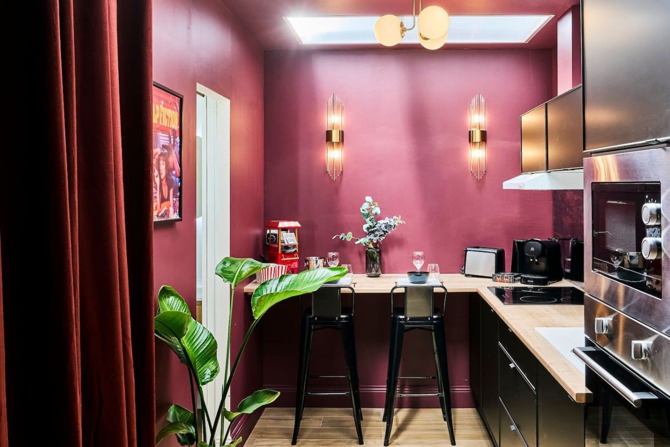 A compact kitchen with a red wall, two bar stools, a coffee machine, a toaster, and a modern refrigerator.