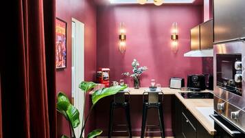 A compact kitchen with a red wall, two bar stools, a coffee machine, a toaster, and a modern refrigerator.