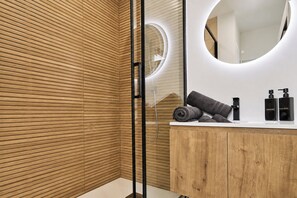 A modern bathroom with wooden paneling, a round mirror, a white vanity, and toiletries. - Vrbo Property (Armentières)