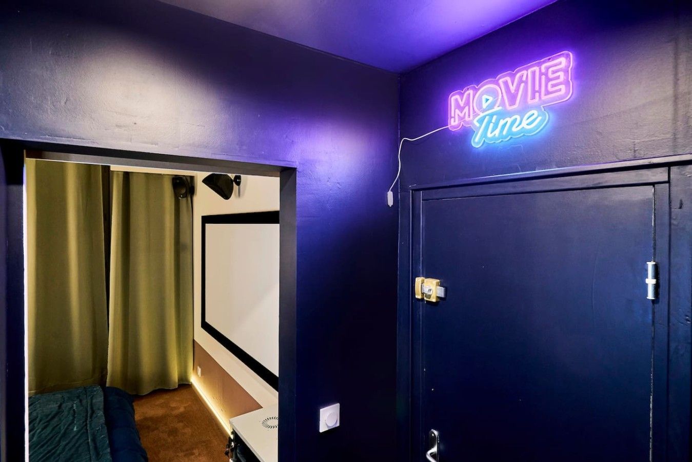 A room with a neon sign reading 'MOVIE Time' on the wall, a door with a handle, and a partial view of a bed with a blanket.