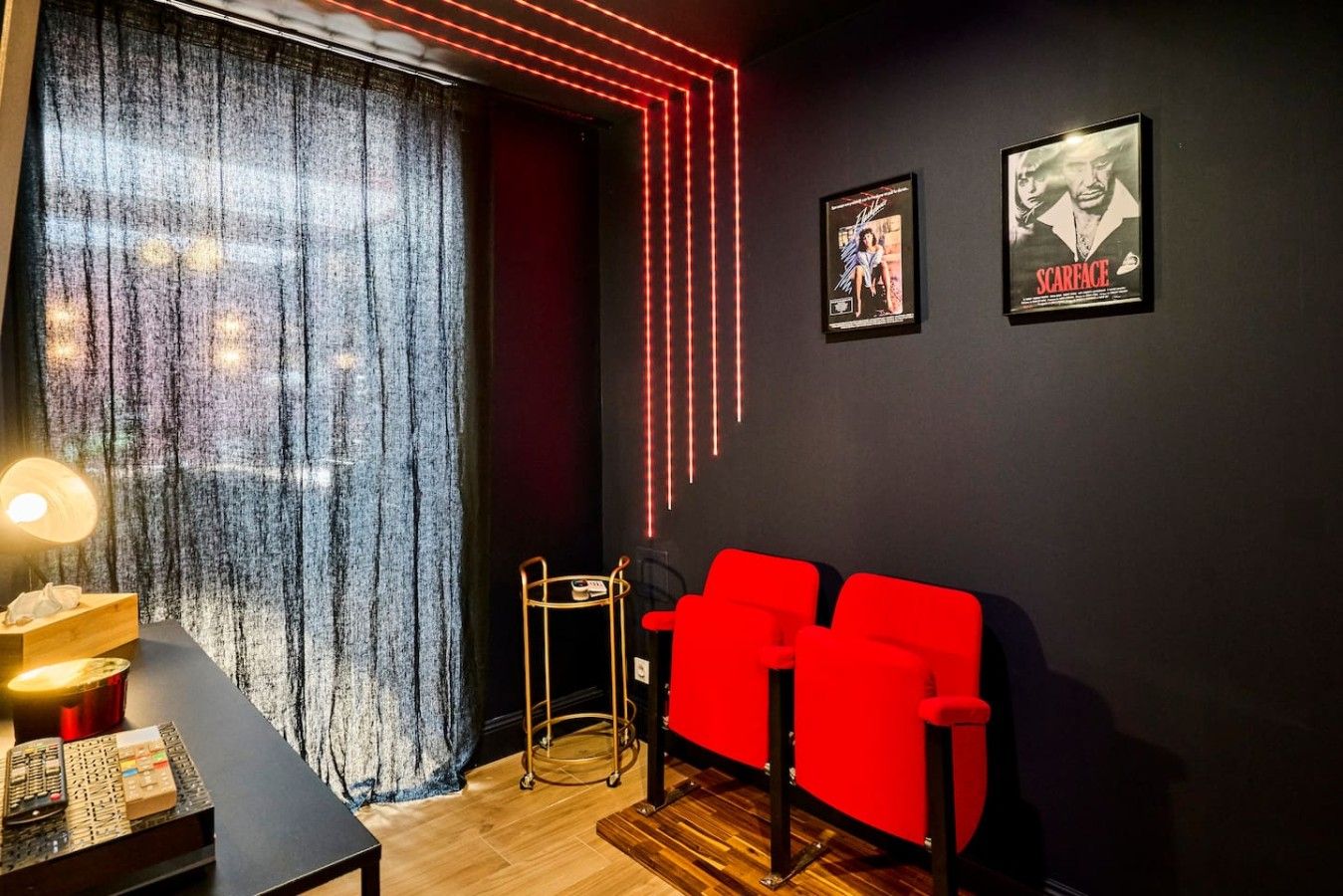 Una stanza con una parete scura, due poster di film incorniciati, un'area salotto rossa, un tavolino con una lampada e una finestra coperta da tende.