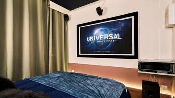 Ein Hotelzimmer mit großem Flachbildfernseher an der Wand, einem Bett mit blauer Bettdecke, einem Deckenprojektor und einer weißen Klimaanlage.
