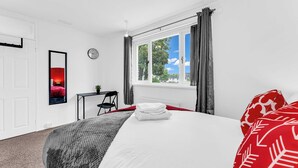 A hotel room with a bed, a desk, a chair, a window with curtains, a clock, and a mirror. - Arcade Machine | Games | Large House | Sleeps 6 (Coventry)