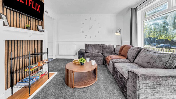 A modern living room with a Netflix sign, a clock on the wall, a sofa, a coffee table, and a window with a view of the outside. - Arcade Machine | Games | Large House | Sleeps 6 (Coventry)