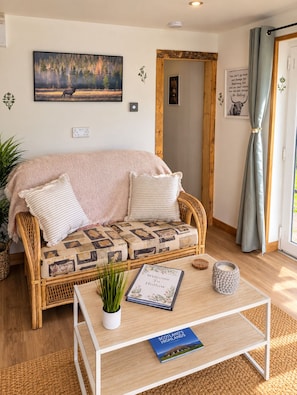 Ein gemütliches Zimmer mit einem Korbsessel, einem kleinen Tisch mit einem Buch und einer Kerze sowie einem Bild eines Rehs an der Wand.
