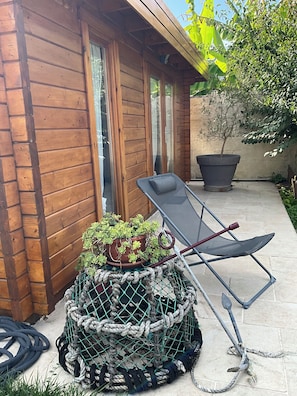 A wooden cabin with a sliding glass door, a reclining chair, a potted plant, and a large planter.