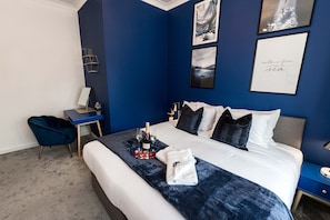 Ein Hotelzimmer mit einem Bett, einem Schreibtisch und einem Sessel. An der Wand hängen zwei gerahmte Bilder, auf dem Bett steht eine Champagnerflasche und ein Tablett mit Gläsern und Erdbeeren.