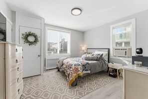 Ein Schlafzimmer mit Bett, Kommode, Nachttisch, Lampe und einem Fenster mit Jalousien.