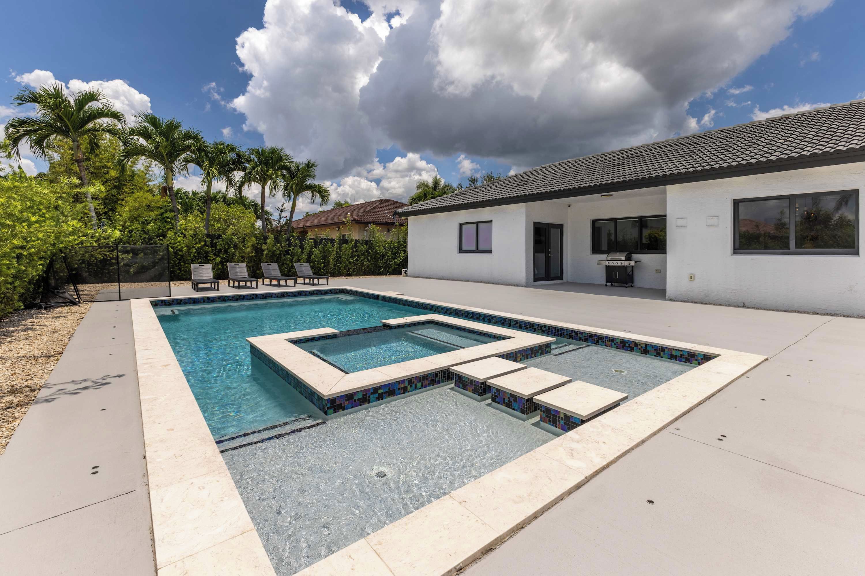 A swimming pool with a geometric design, surrounded by a patio area, palm trees, and a house with a tiled roof.