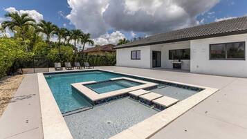 A swimming pool with a geometric design, surrounded by a patio area, palm trees, and a house with a tiled roof.