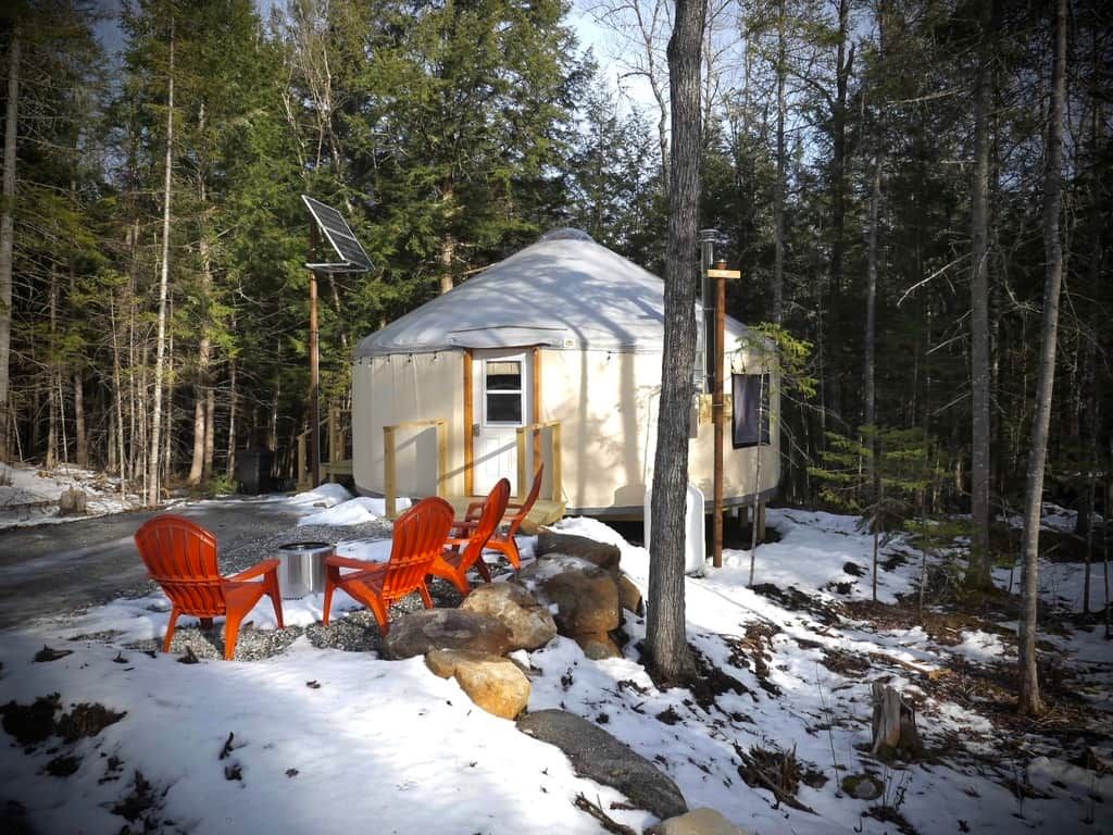 En lille hytte med en dør og et vindue, omgivet af skov. To orange stole og et lille bord står udenfor.
