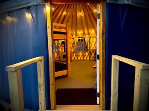 A wooden interior of a yurt with a central sleeping area and a door leading outside.