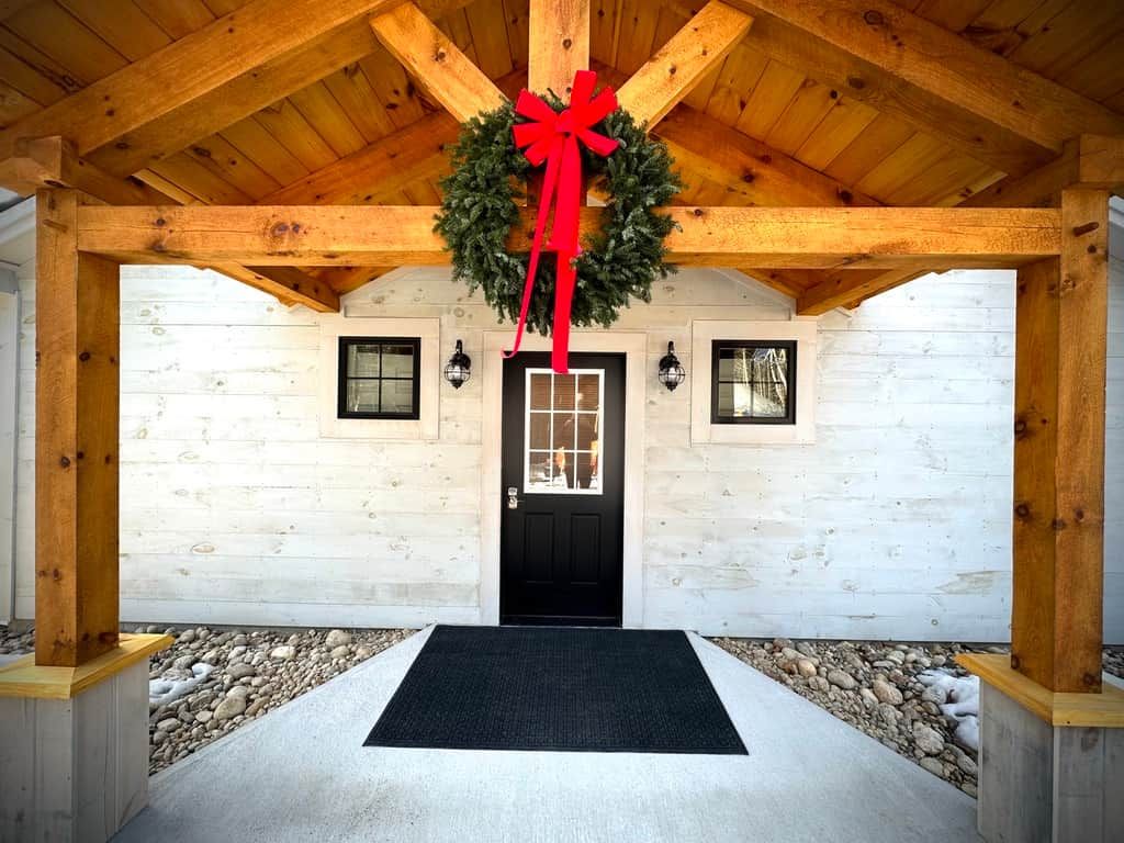 A wooden exterior with a black door, a red bow on a wreath, and a concrete pathway leading to the door.