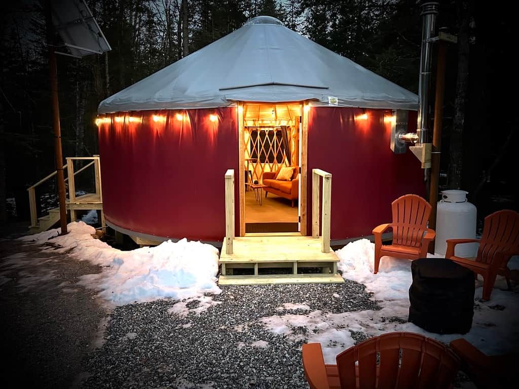 A yurt with a warm interior light, a wooden entrance, and outdoor seating.