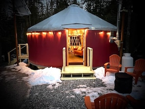 A yurt with a warm interior light, a wooden entrance, and outdoor seating.