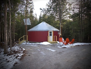 Una yurta roja con puerta y ventanas, rodeada por un bosque, con un camino de grava que la lleva a ella y dos sillas naranjas afuera.