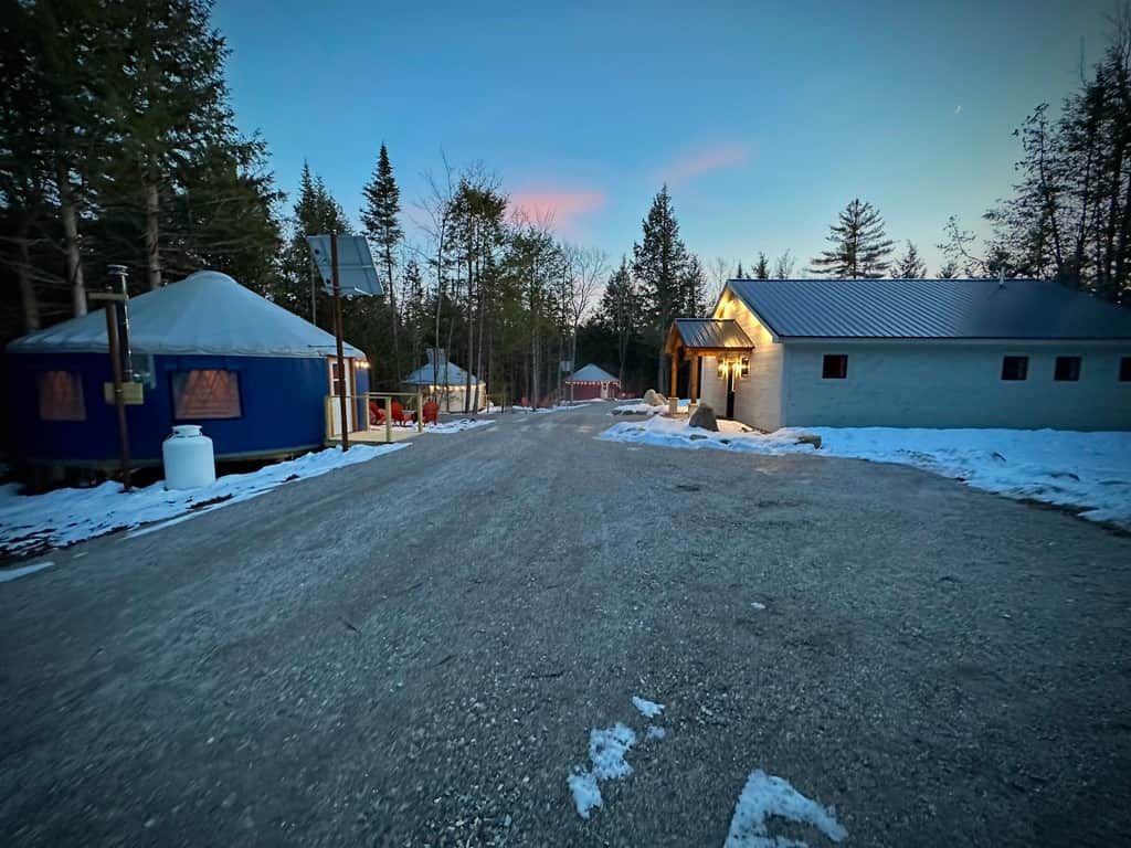 Maaseututie, jonka varrella on jurttamajoja ja pieni rakennus, ja kaikkialla ympärillä on puita.