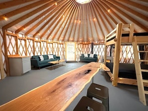 A yurt interior with a central wooden table, two sofas, a bunk bed, and a small kitchenette.