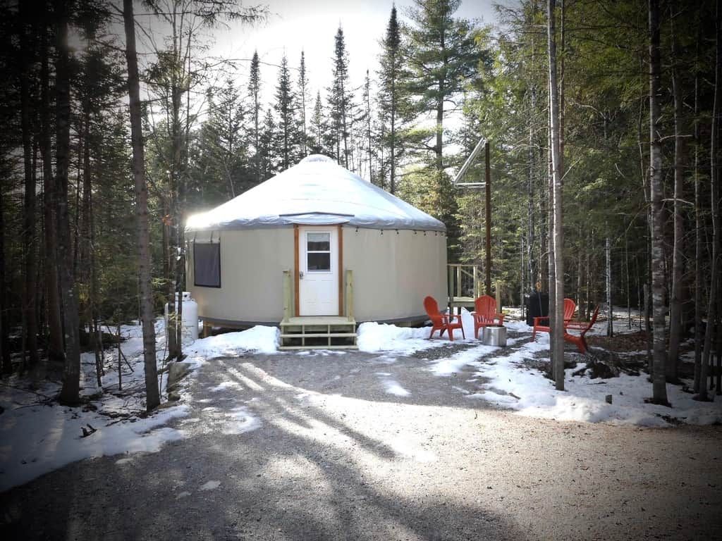 A yurt with a door and steps, surrounded by trees and snow, with two red chairs outside.