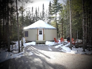 A yurt with a door and steps, surrounded by trees and snow, with two red chairs outside.