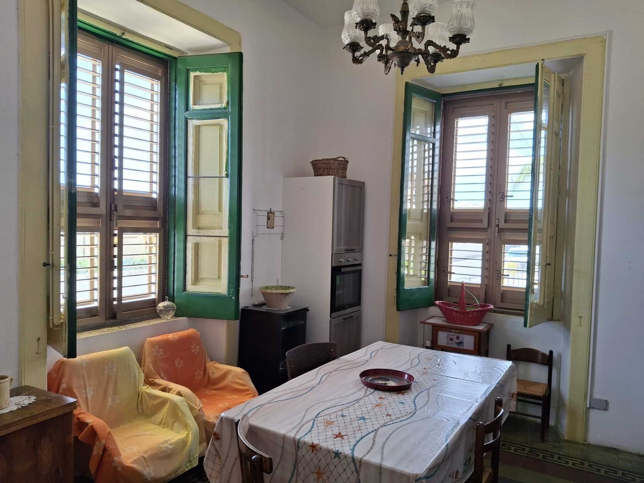 Ein Zimmer mit Esstisch, Stühlen, einer Kandelaber und Fenstern mit Fensterläden.