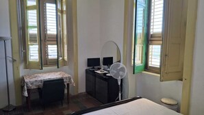 Quarto com cama, escrivaninha, cadeira, ventilador e duas janelas com persianas abertas.