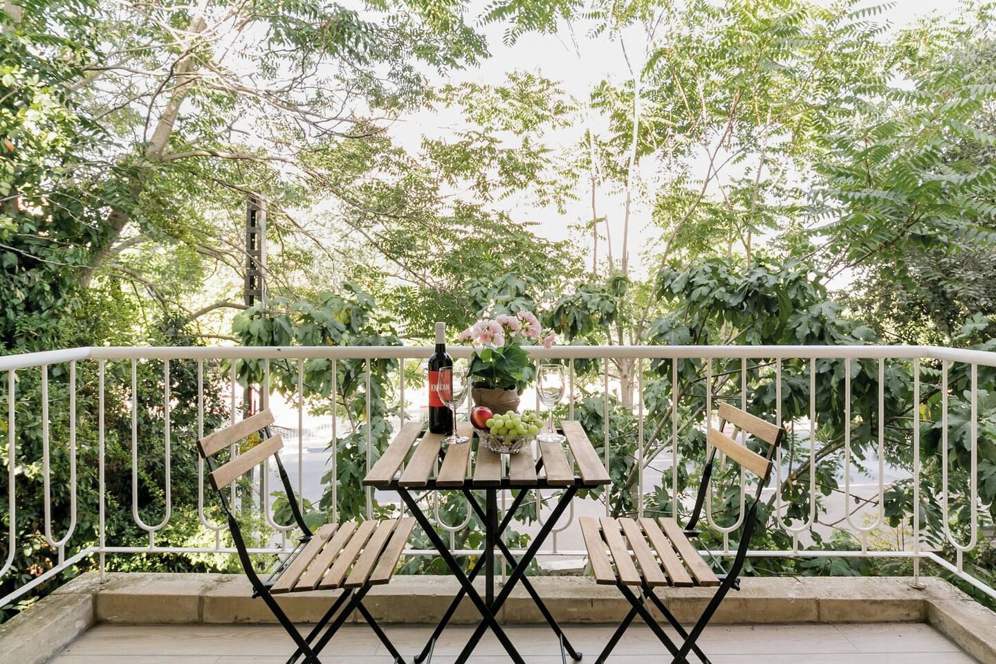 An outdoor balcony with a wooden table, two chairs, a bottle of wine, a glass, and a bowl of fruit.