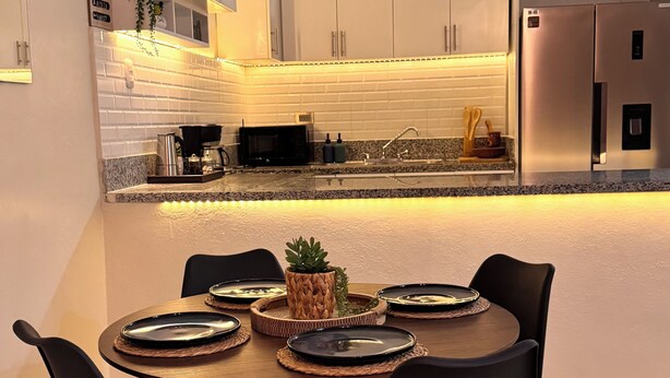 A modern kitchen with white cabinets, a central island with a sink, and a dining area with a round table and chairs. - 3-Bedroom Apartment 20 Minutes from AILA and the Colonial District (Santo Domingo Este)
