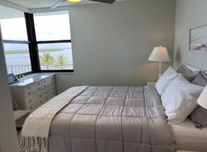 A bedroom with a bed, a nightstand, a lamp, a dresser, and a window with a view of the water. - One Bedroom Condo with amazing Bayviews #504 (Fort Myers Beach)