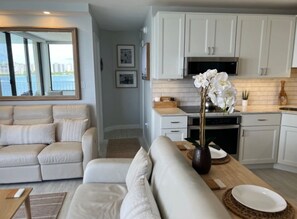 A modern living room with a sofa, a dining area with a table and chairs, and a kitchen with white cabinets and a microwave. - One Bedroom Condo with amazing Bayviews #504 (Fort Myers Beach)