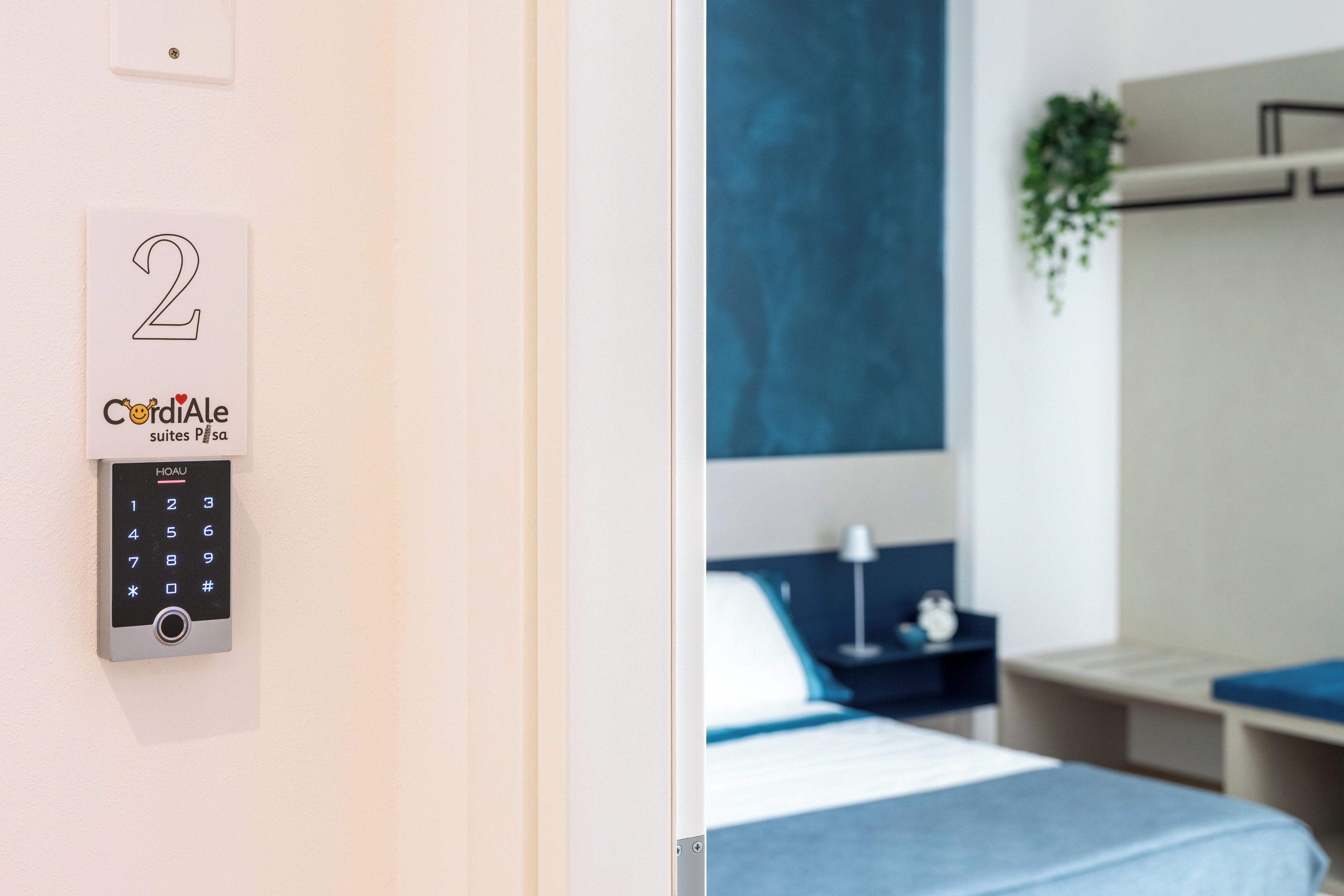 Habitación de hotel con cerradura electrónica de teclado numérico, cama con cabecera azul, mesita de noche con lámpara y planta en la pared.