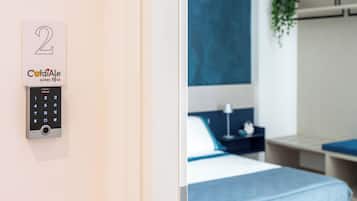 Habitación de hotel con cerradura electrónica de teclado numérico, cama con cabecera azul, mesita de noche con lámpara y planta en la pared.