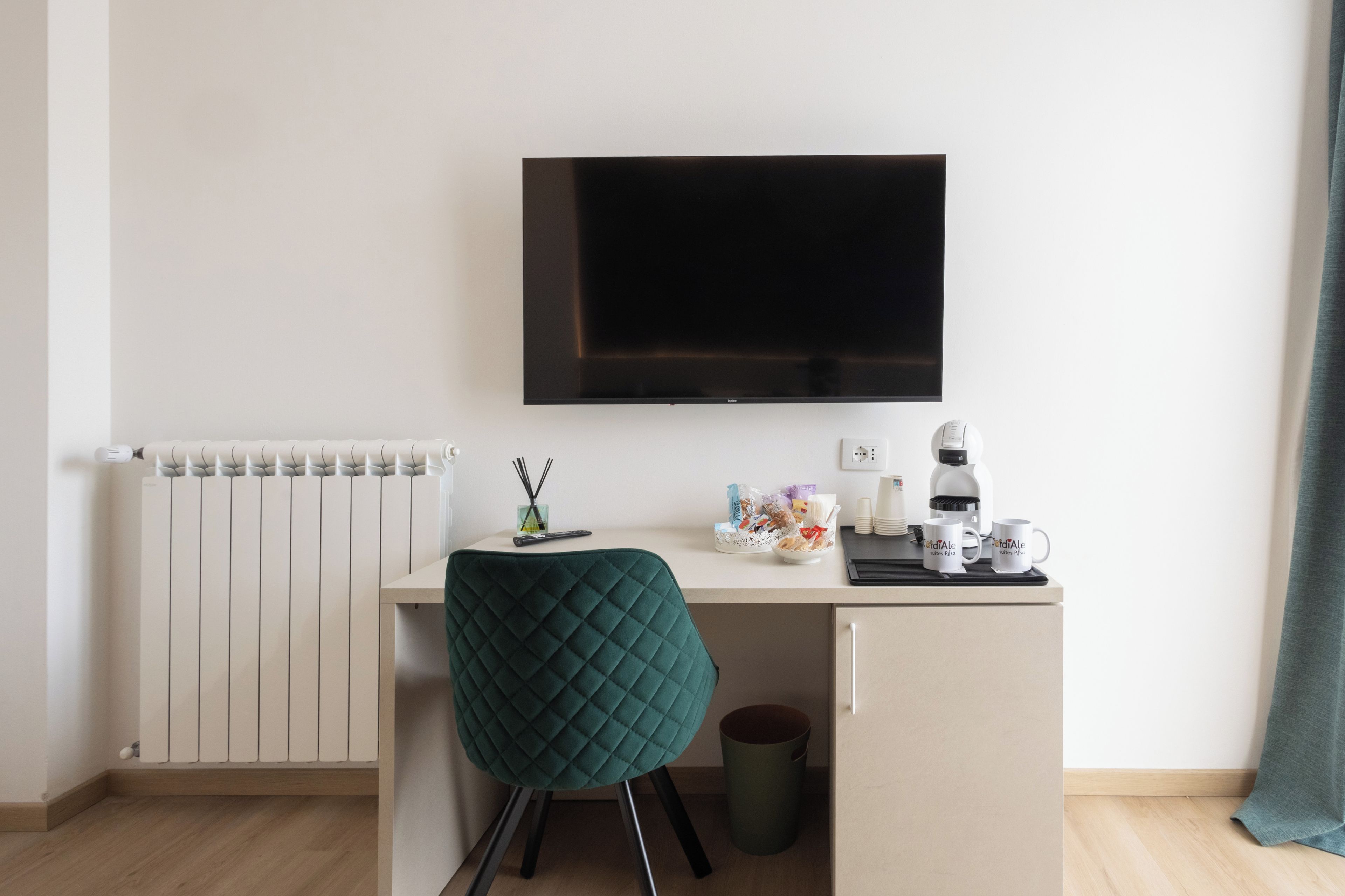 A modern, minimalistic room with a flat-screen TV mounted on the wall, a radiator, a desk with a green chair, a coffee machine, and a trash bin.
