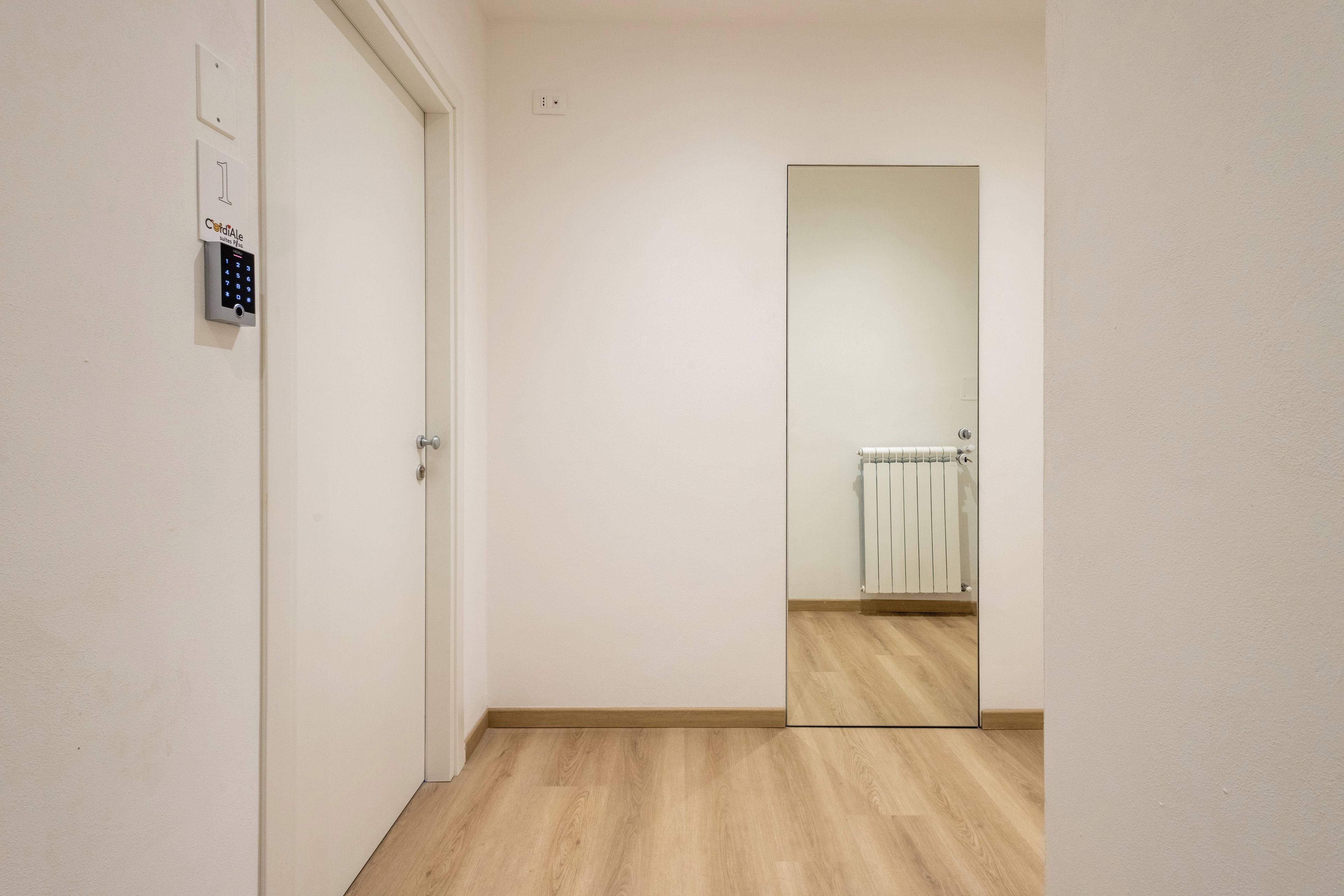 Een gang met een deur, een beveiligingspaneel aan de muur en een grote spiegel die een radiator weerspiegelt.