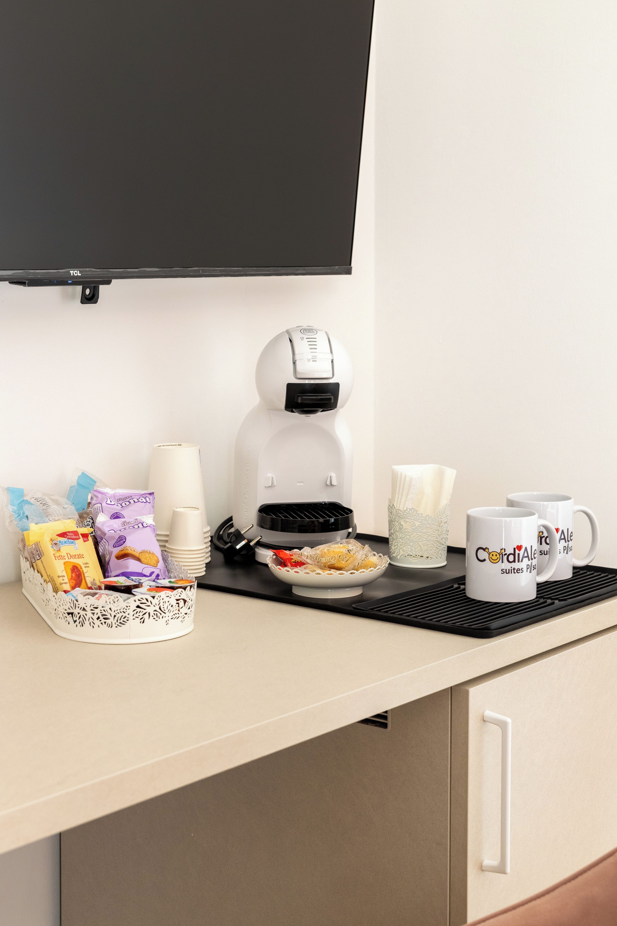 A hotel room's breakfast setup with a coffee maker, a plate of pastries, a tray with condiments, and a mug with a heart design.
