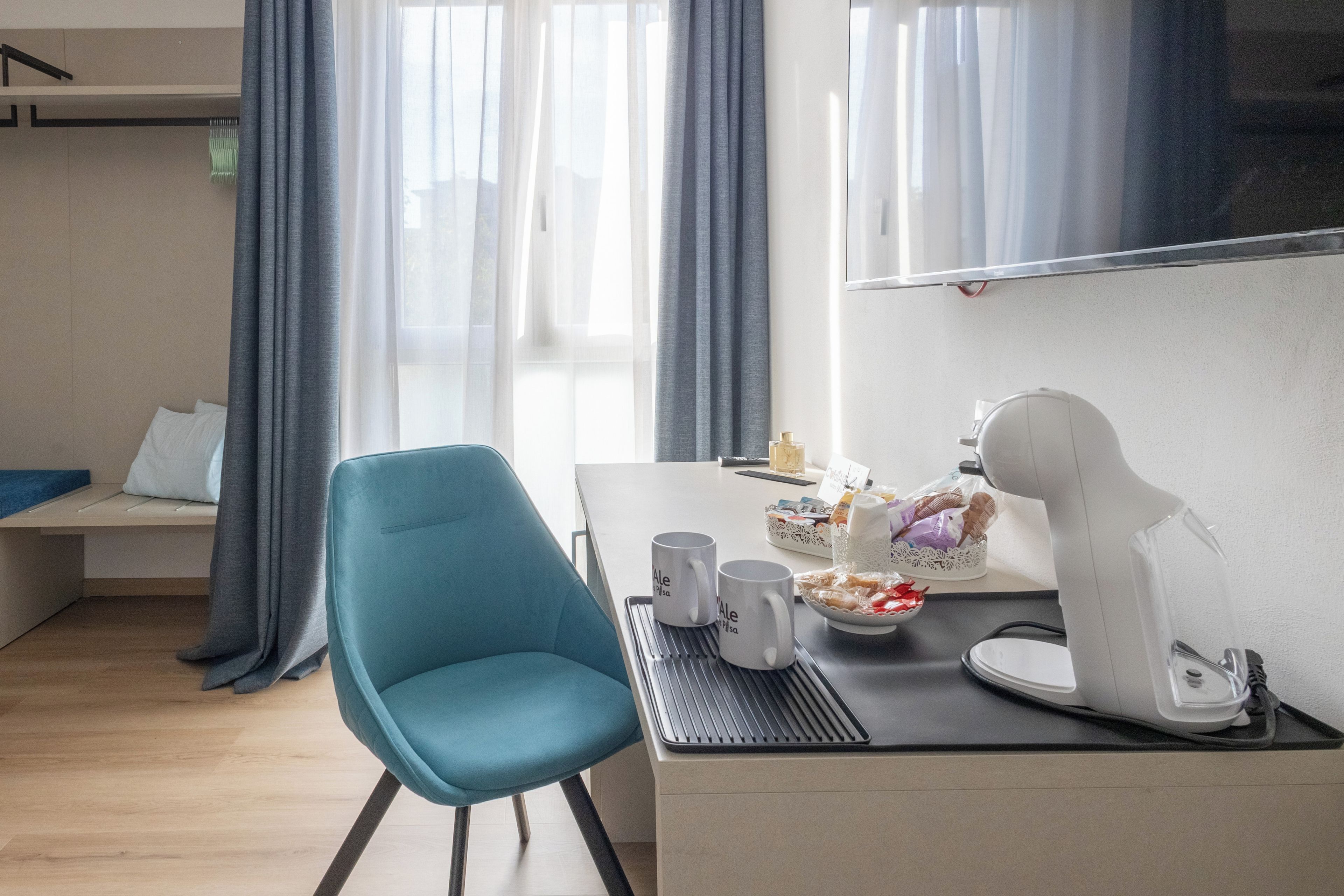 Ein modernes Zimmer mit einem türkisgrünen Sessel, einem Schreibtisch mit einem Laptop, einer Kaffeemaschine und einer Auswahl an Snacks.