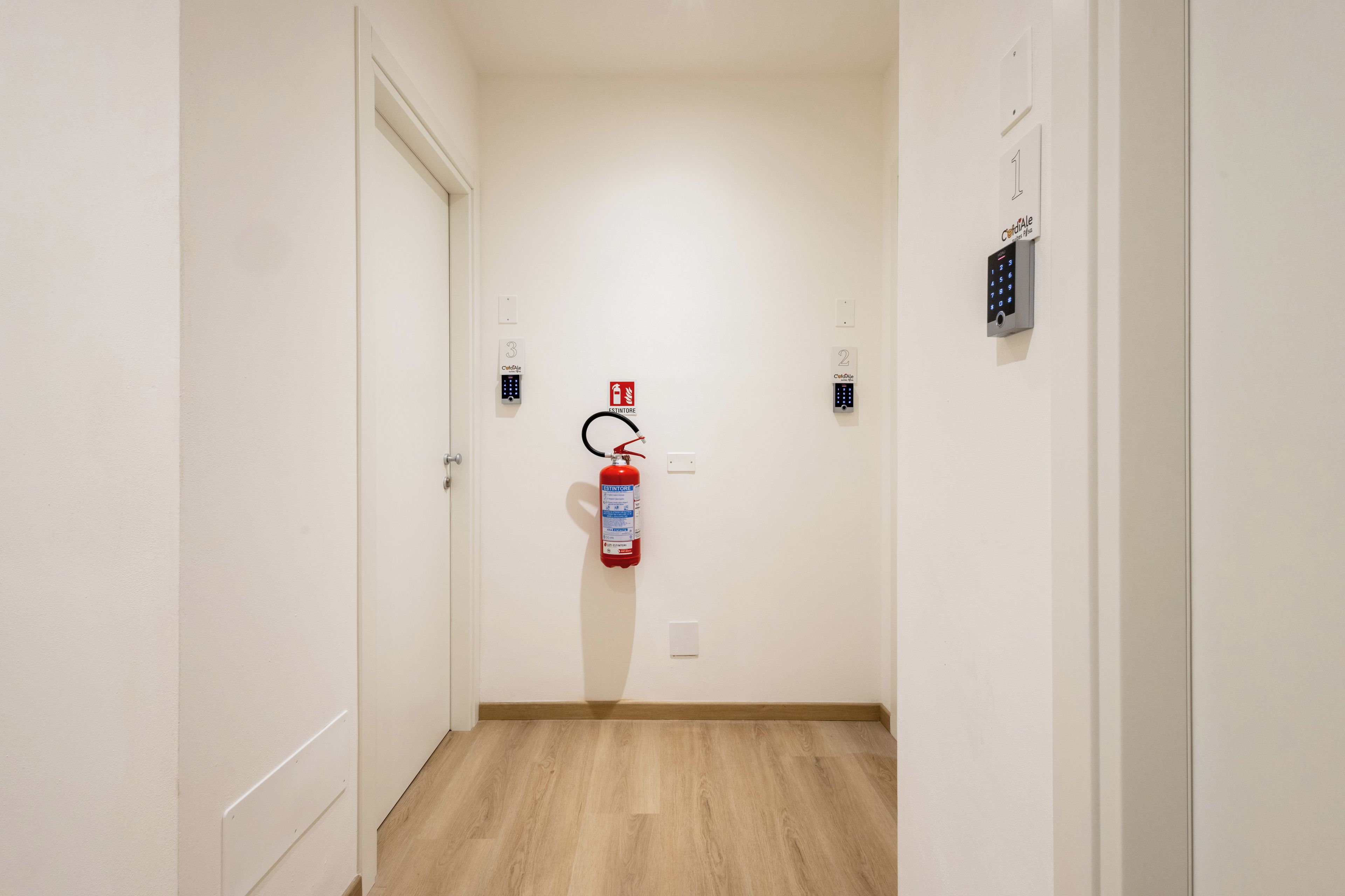 A hallway with a fire extinguisher mounted on the wall, a door at the end, and a keypad on the right wall.
