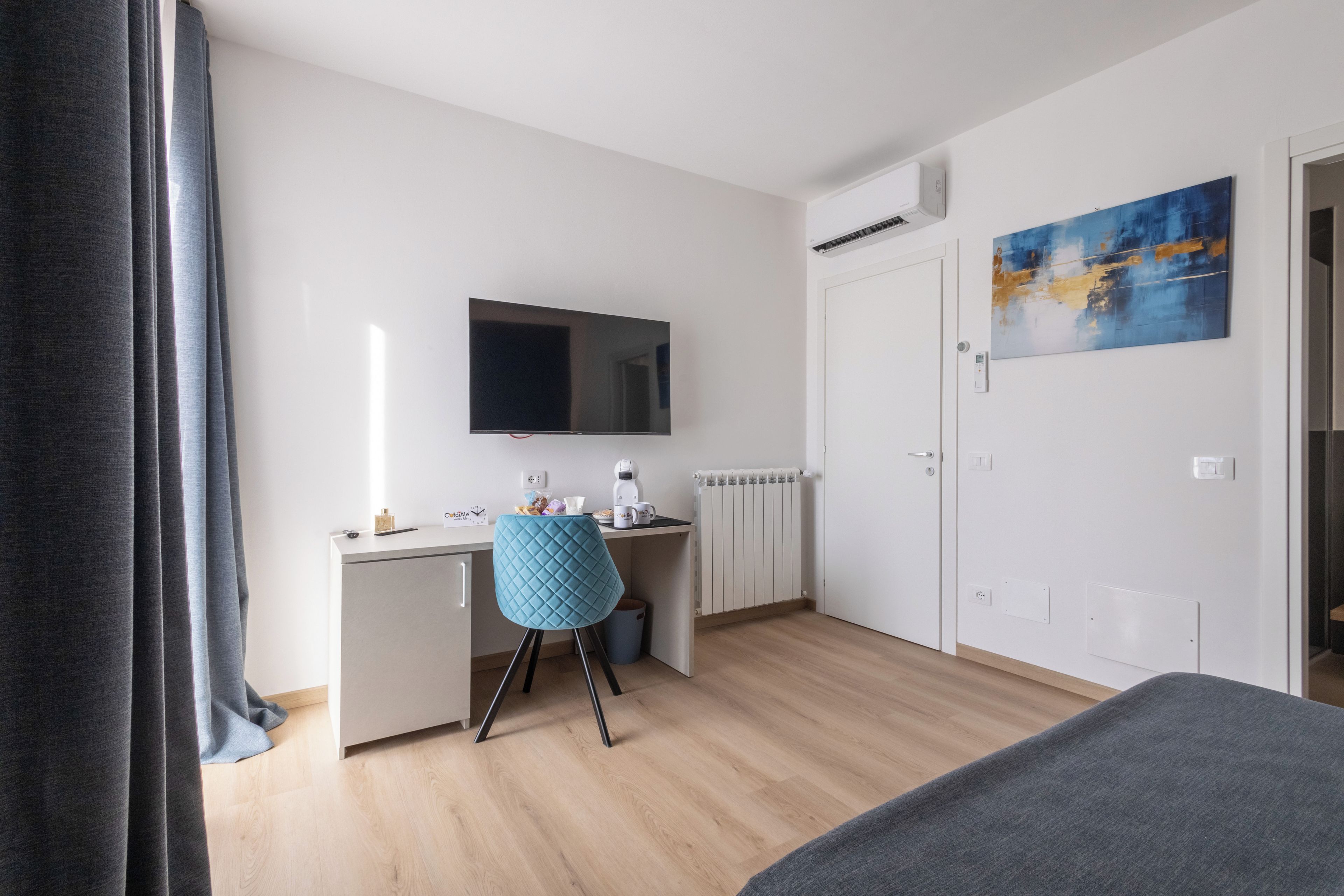 A modern hotel room with a flat-screen TV, a desk with a chair, a radiator, a door with a painting, and a window with curtains.