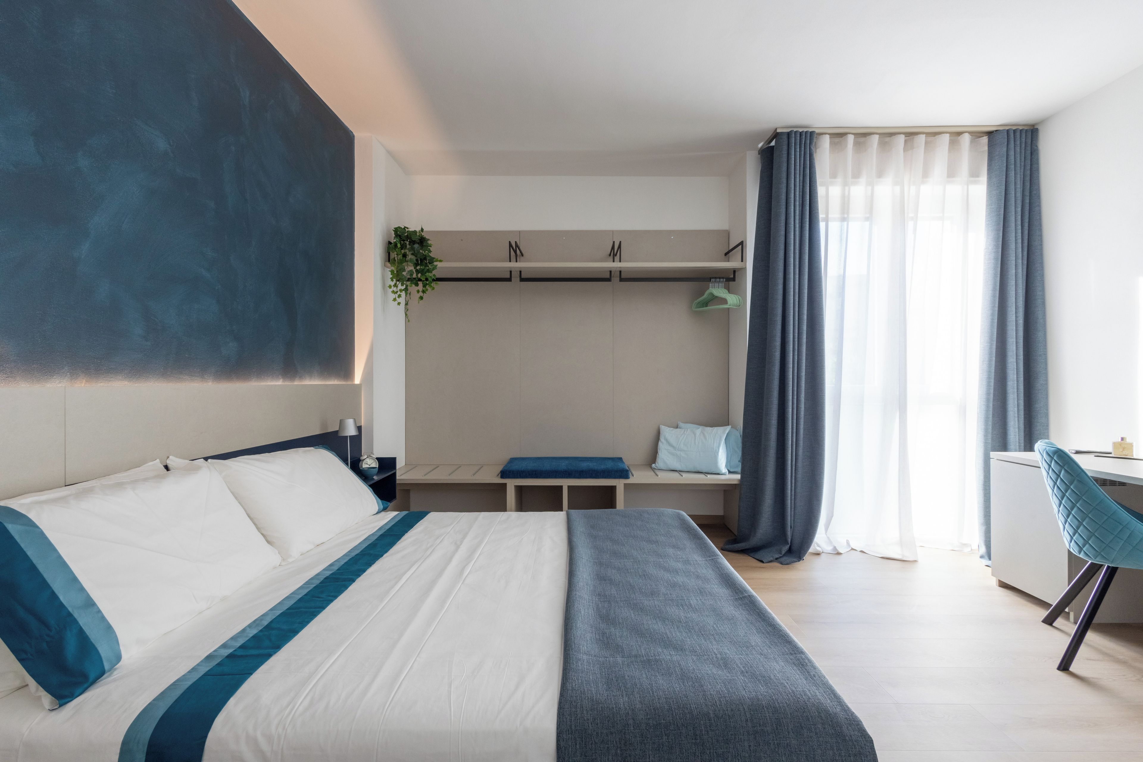 Una habitación de hotel con cama, escritorio, silla, armario y ventana con cortinas.