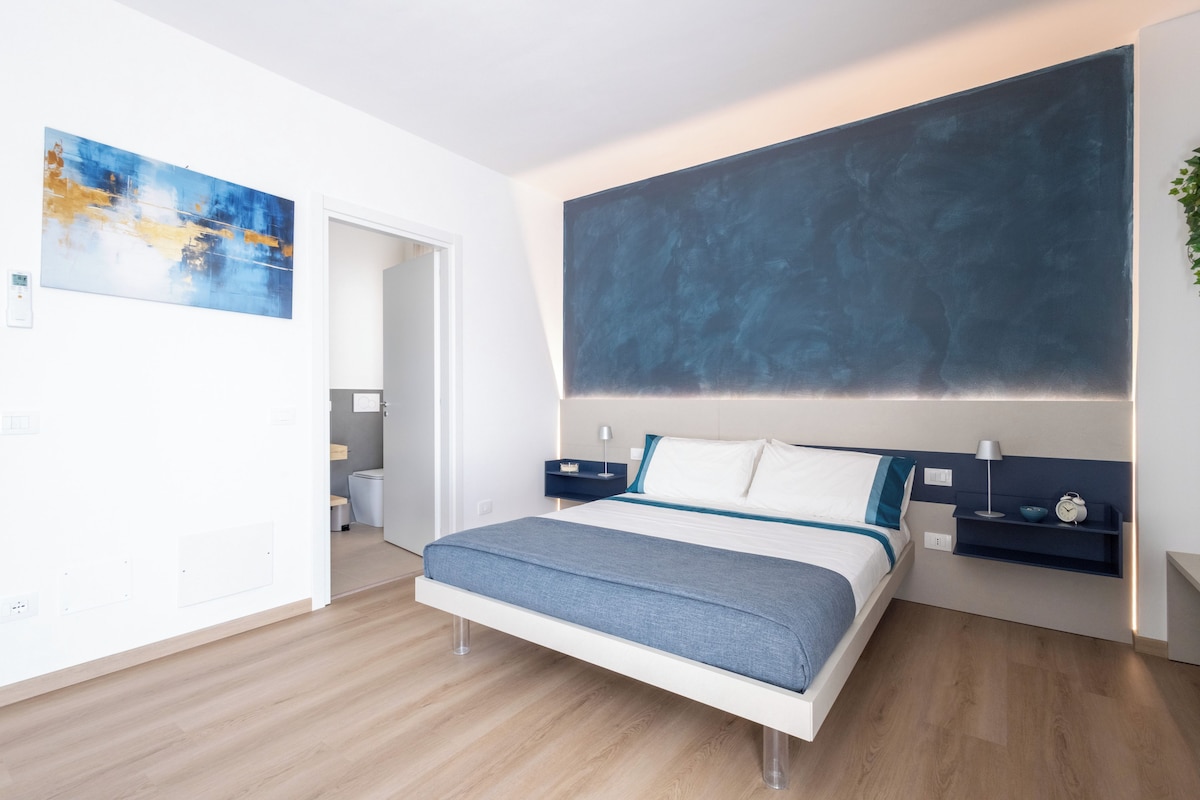 A modern bedroom with a bed, bedside tables, a large abstract painting, and a view into the bathroom.