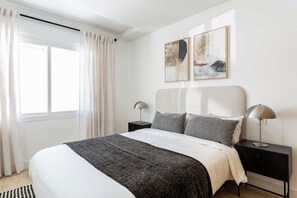 Ein modernes Schlafzimmer mit einem großen Bett, zwei Nachttischen, einem Fenster mit Vorhängen und abstrakter Wandkunst.