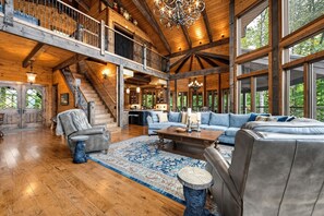 Un intérieur spacieux avec de hauts plafonds, des poutres en bois, un escalier, un coin salon avec des canapés et une table basse, et de grandes fenêtres offrant une vue sur l’extérieur.
