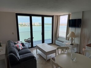 A living room with a sofa, a table, and a chair, a balcony with a view of the sea, and a TV mounted on the wall. - One Bedroom Condo with stunning Water Views #1103 (Fort Myers Beach)