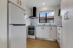 A compact kitchen with white cabinetry, a built-in oven, and a window with a view of a red-roofed building. - Lake & Tide: A fresh, modern escape close to Cunninghame Arm (Lakes Entrance)
