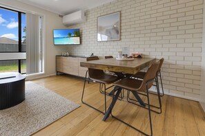 A modern kitchen with a wooden table and chairs, a TV mounted on the wall, and a brick-patterned backsplash. - Lake & Tide: A fresh, modern escape close to Cunninghame Arm (Lakes Entrance)
