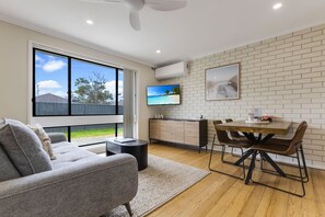 A modern living room with a grey sofa, a dining table with chairs, a TV on the wall, and a large window with a view of the outdoors. - Lake & Tide: A fresh, modern escape close to Cunninghame Arm (Lakes Entrance)