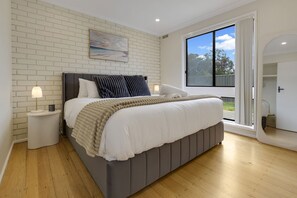 Un dormitorio moderno con una cama tapizada en gris, ropa de cama blanca, una pared de ladrillo blanco, un ventanal con vistas y una mesita de noche pequeña con una lámpara.